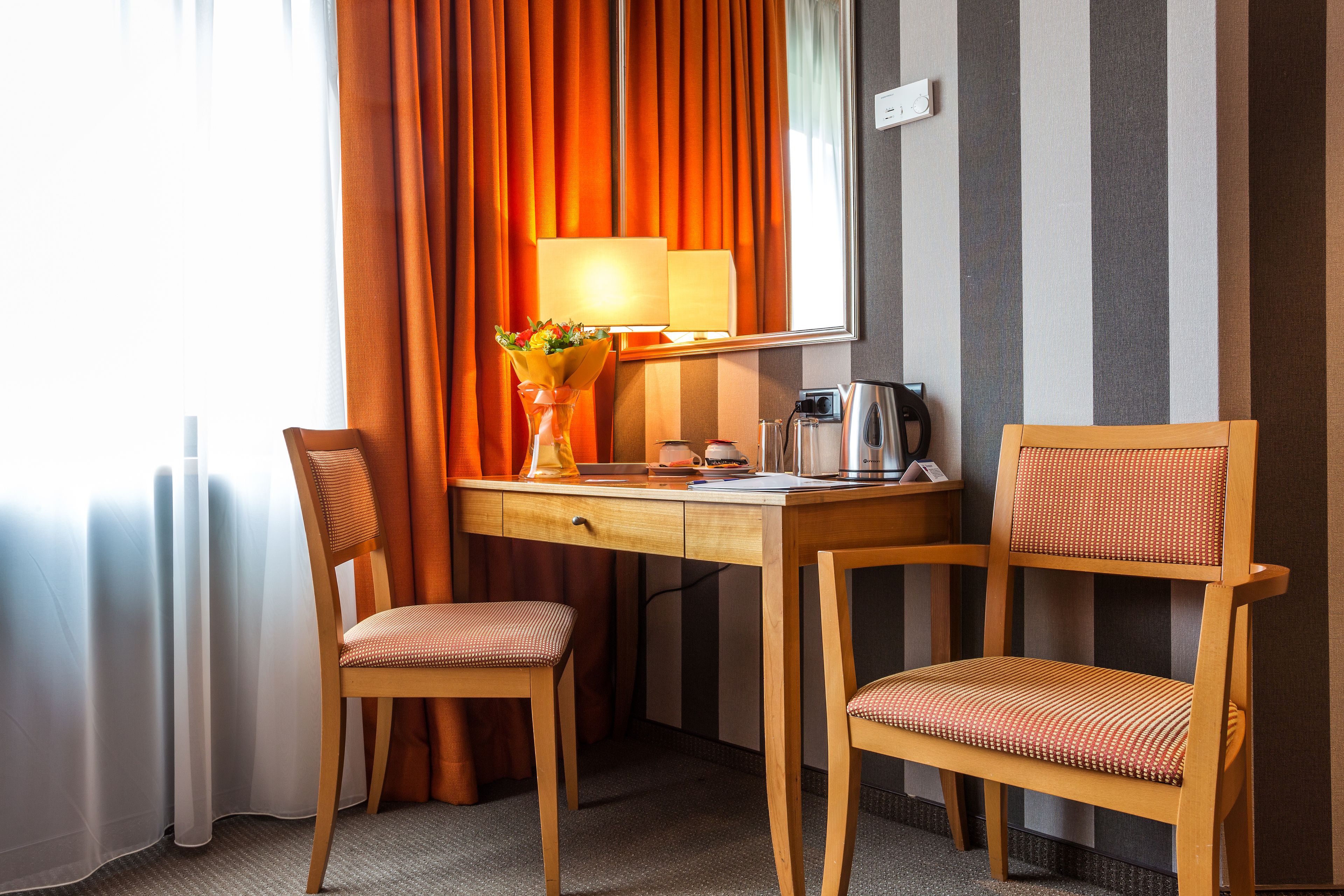 Een hotelkamer met een houten bureau, twee stoelen, een lamp, een koffiezetapparaat en een raam met oranje gordijnen.