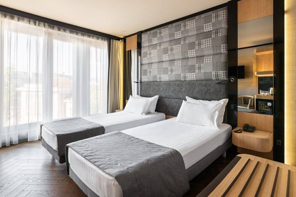 Een hotelkamer met twee bedden, een houten bankje, een nachtkastje met een telefoon en een groot raam met dunne gordijnen.