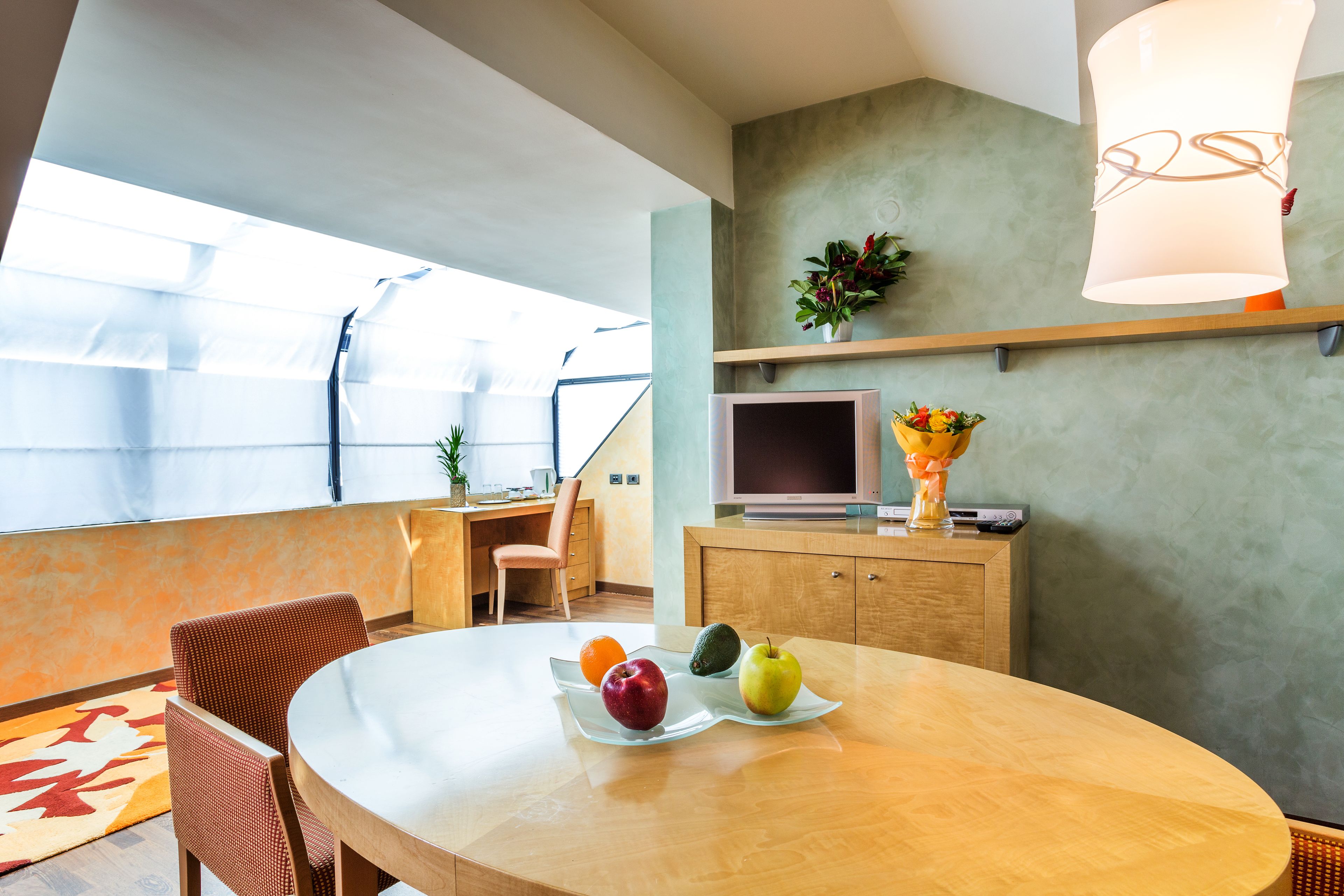 Una sala moderna con una mesa redonda de madera, un televisor y un sofá. Hay dos sillones y una pequeña planta en una estantería.