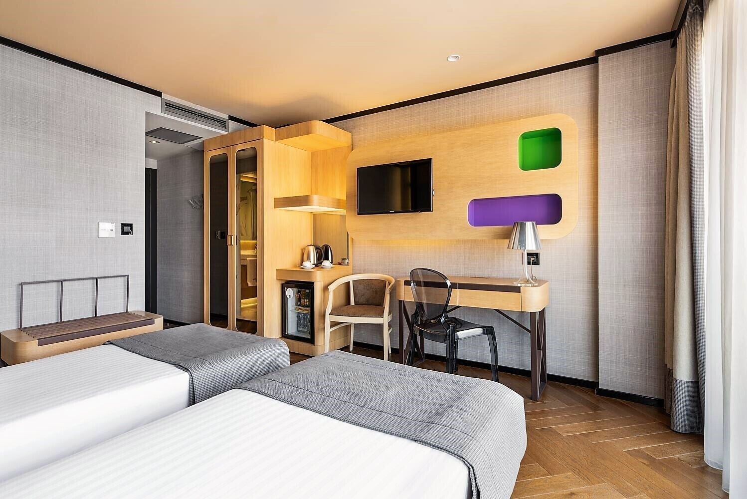A hotel room with two beds, a desk, a chair, a television, and a minibar.