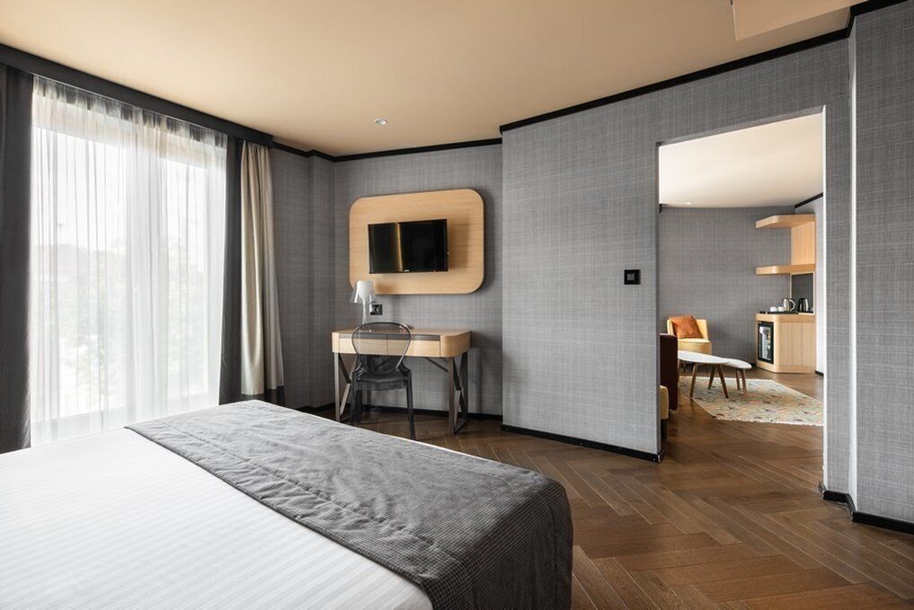 A modern hotel room with a large bed, a desk with a chair, a television mounted on the wall, and a view of the city through the window.