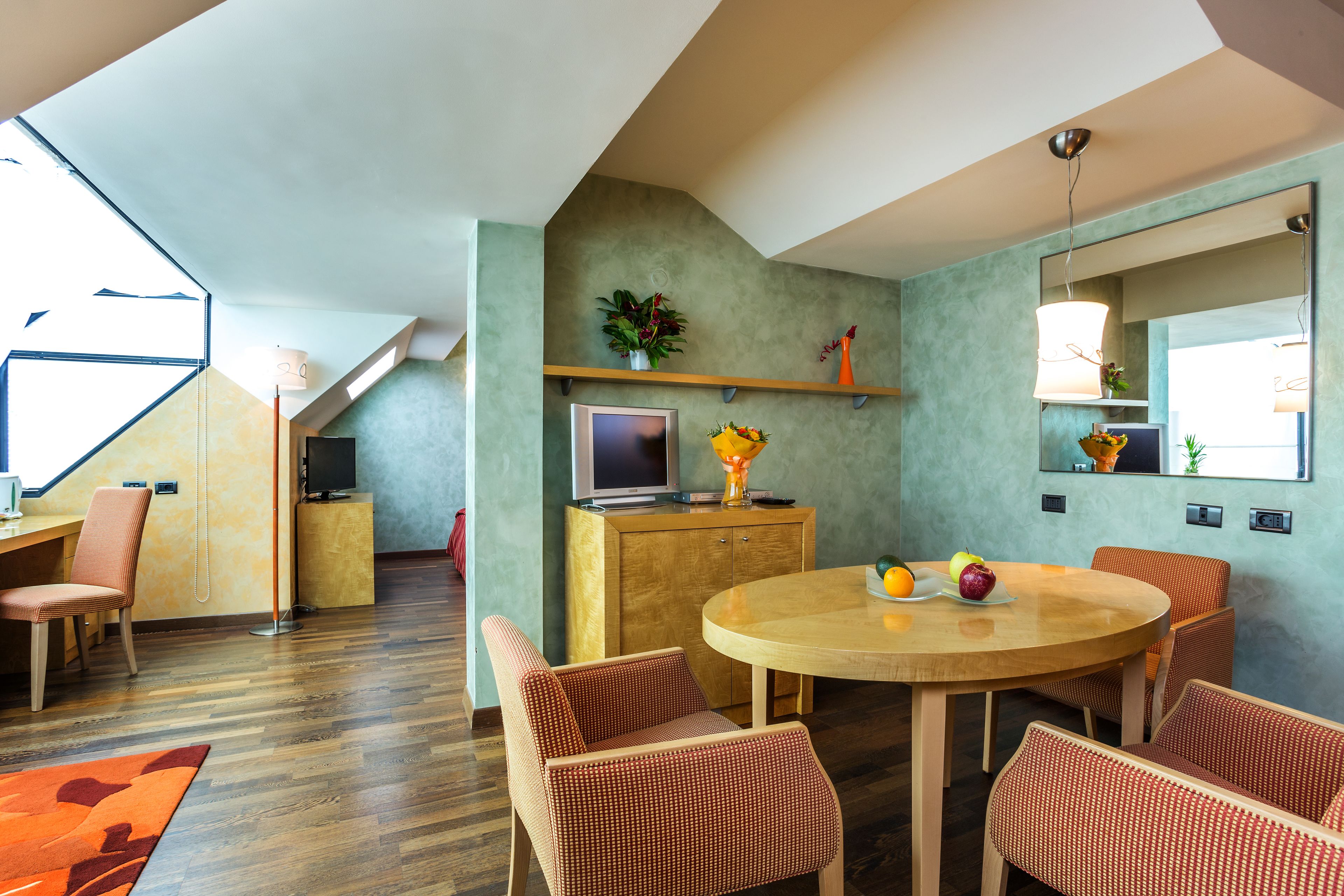 Un salon moderne avec une table ronde en bois, des chaises à motifs rouges, un téléviseur posé sur un meuble en bois et un grand miroir accroché au mur.
