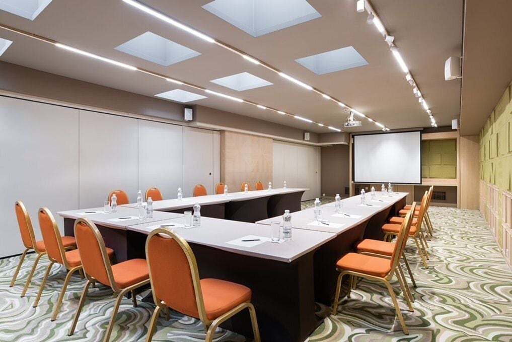 A conference room with a long table, orange chairs, a projector screen, and a patterned carpet.