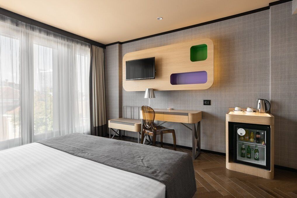 Una habitación de hotel moderna con una cama grande, un escritorio con silla, un televisor montado en un panel de madera y un minibar.