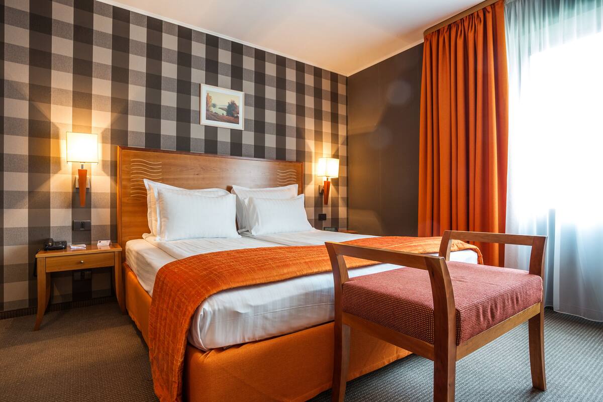 Een hotelkamer met een zwart-wit geruit wandpaneel, een bed met witte lakens en een oranje deken, twee bedlampen, en een houten stoel met een rode kussen.