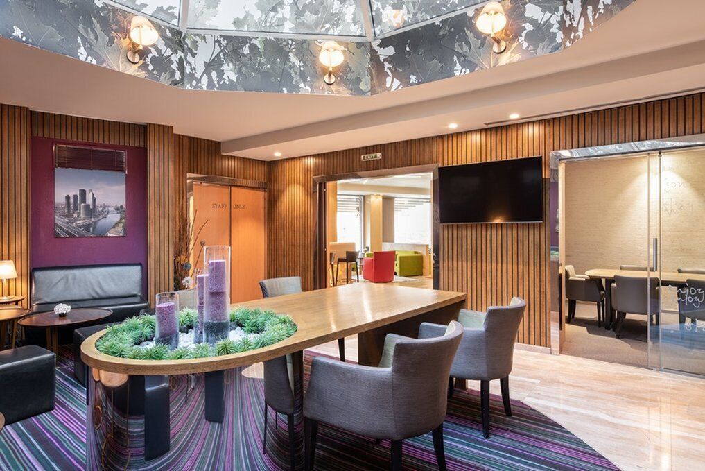 A modern hotel lobby with a large wooden table, comfortable seating, a flat-screen TV, and a cityscape painting on the wall.
