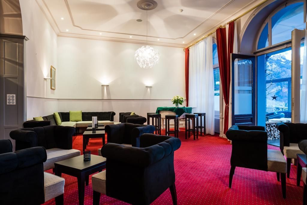 Eine Hotelhalle mit rotem Teppich, einer Sitzecke mit Sofas und Sesseln, einem Kronleuchter und großen, mit Vorhängen versehenen Fenstern.