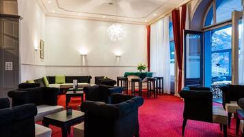 Eine Hotelhalle mit rotem Teppich, einer Sitzecke mit Sofas und Sesseln, einem Kronleuchter und großen, mit Vorhängen versehenen Fenstern.