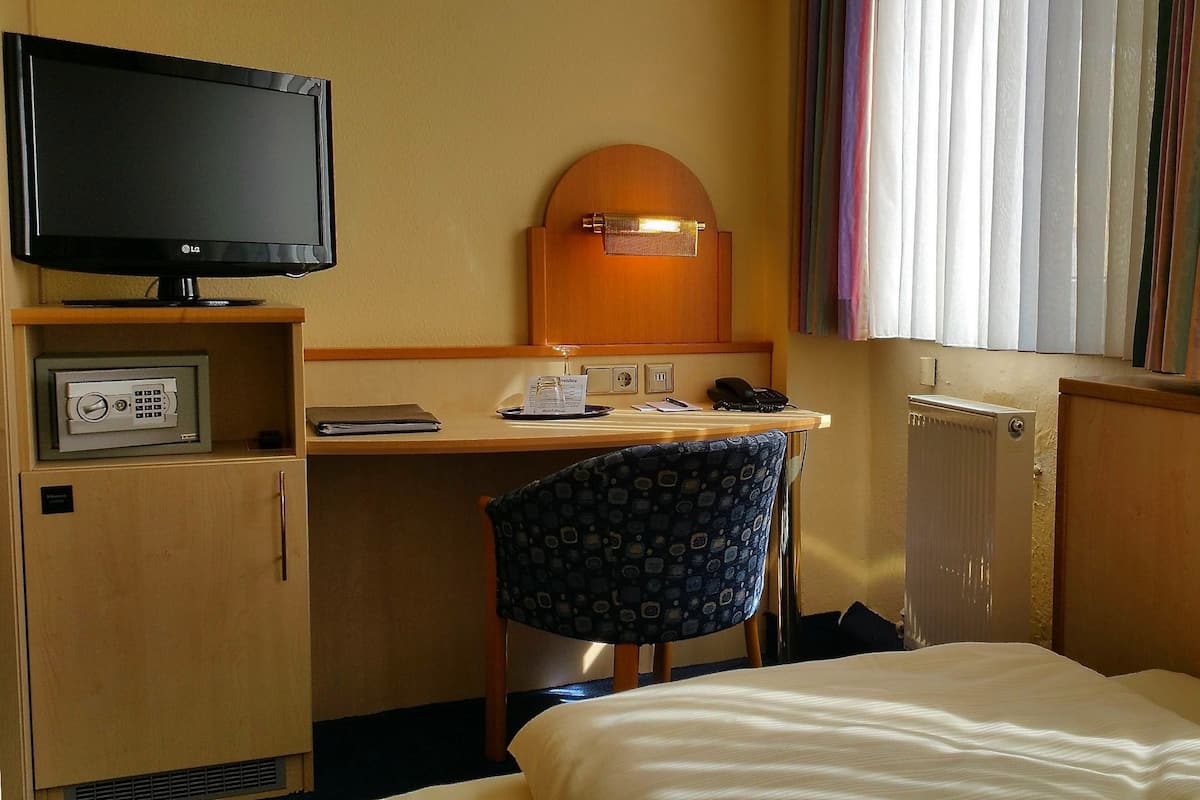 Une chambre d’hôtel comprenant un lit, un bureau avec un ordinateur, une chaise, une télévision et une fenêtre avec des stores.