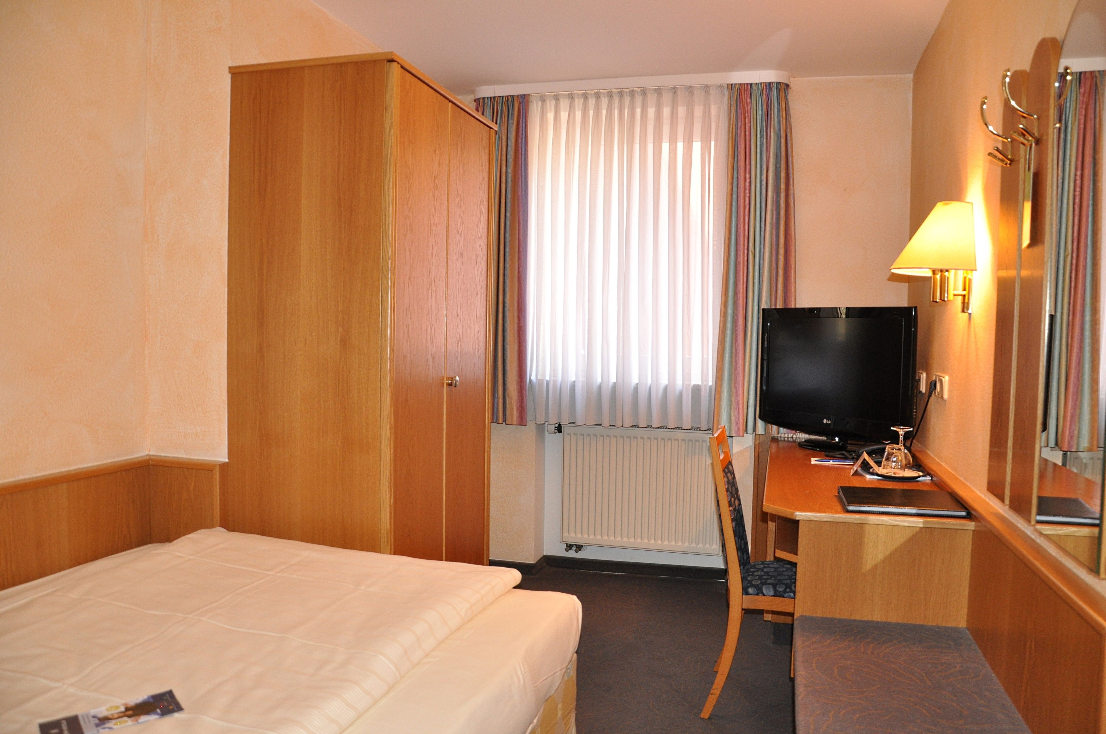 Une chambre d’hôtel avec un lit, un bureau avec une télévision, une chaise et une fenêtre avec des rideaux.