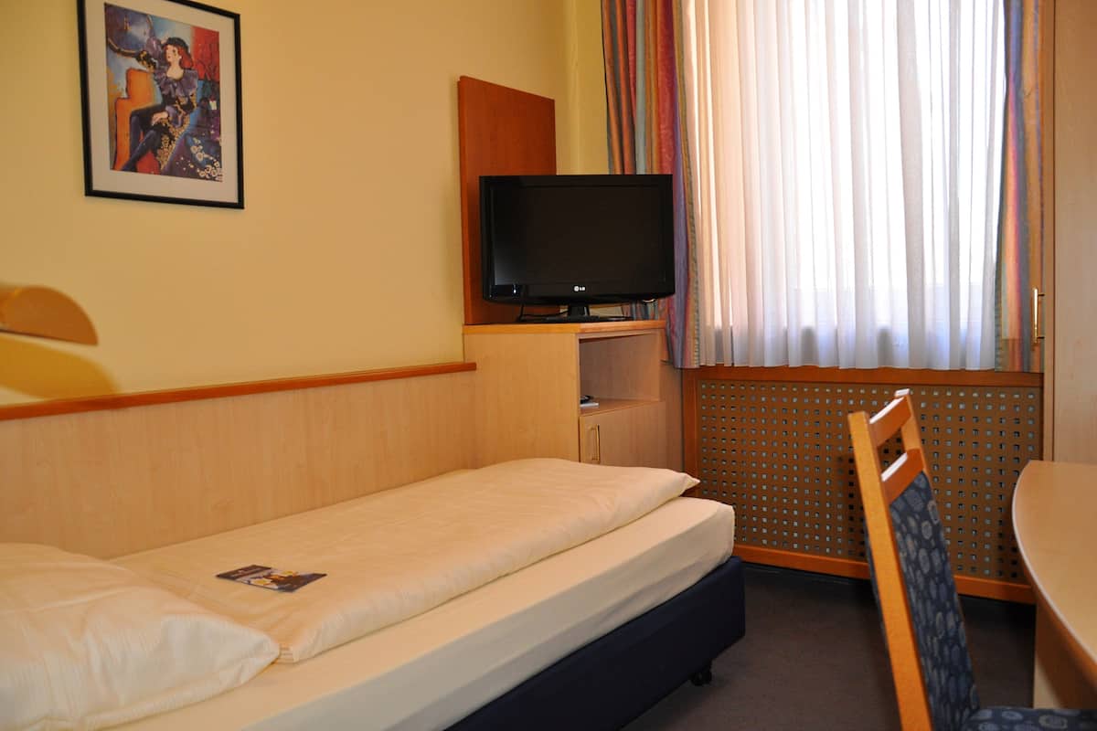 Une chambre d’hôtel avec un lit simple, un téléviseur posé sur un meuble en bois, une petite table et une chaise.