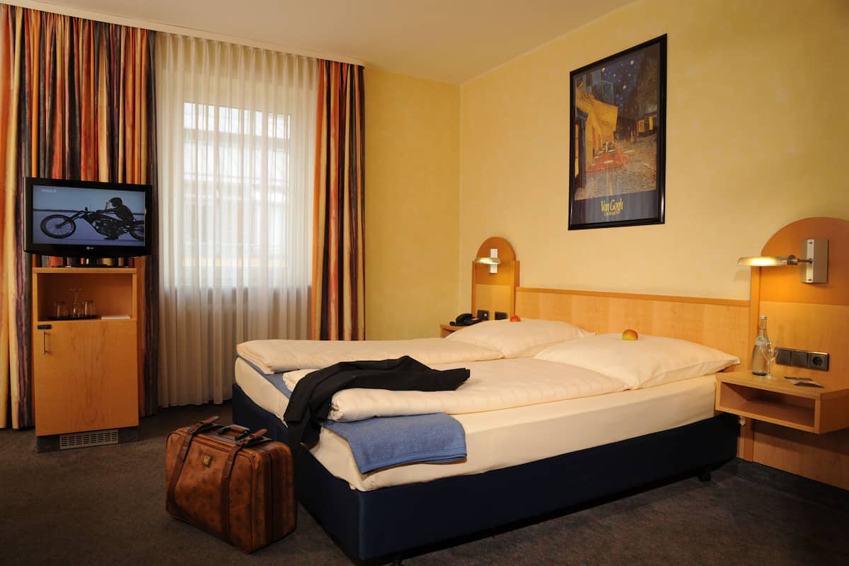 Une chambre d’hôtel avec un lit, une télévision, une table de chevet en bois, une lampe et une valise.