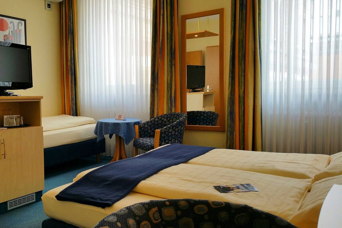 Une chambre d’hôtel comprenant un lit, un bureau avec une chaise, une télévision et une fenêtre avec des rideaux.