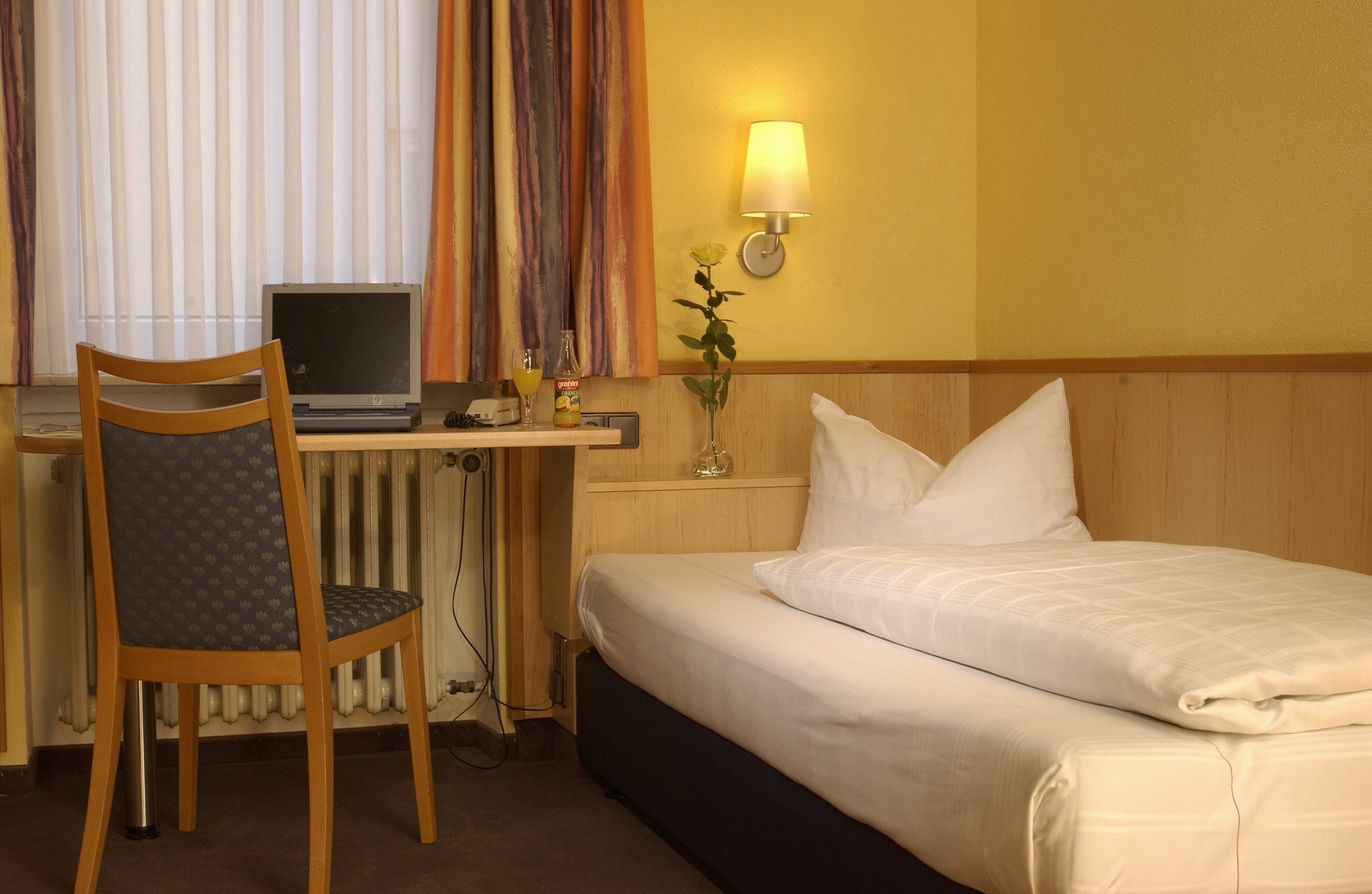 Une chambre d’hôtel avec un lit, un bureau avec un ordinateur portable, une chaise, une lampe et un vase avec des fleurs.