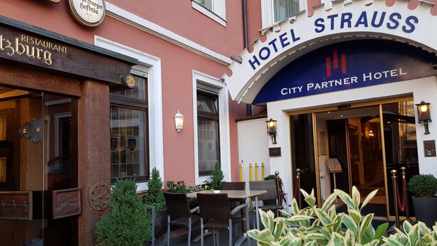 L’entrée d’un hôtel, avec une enseigne indiquant : « Hôtel Strauss City, hôtel partenaire ».
