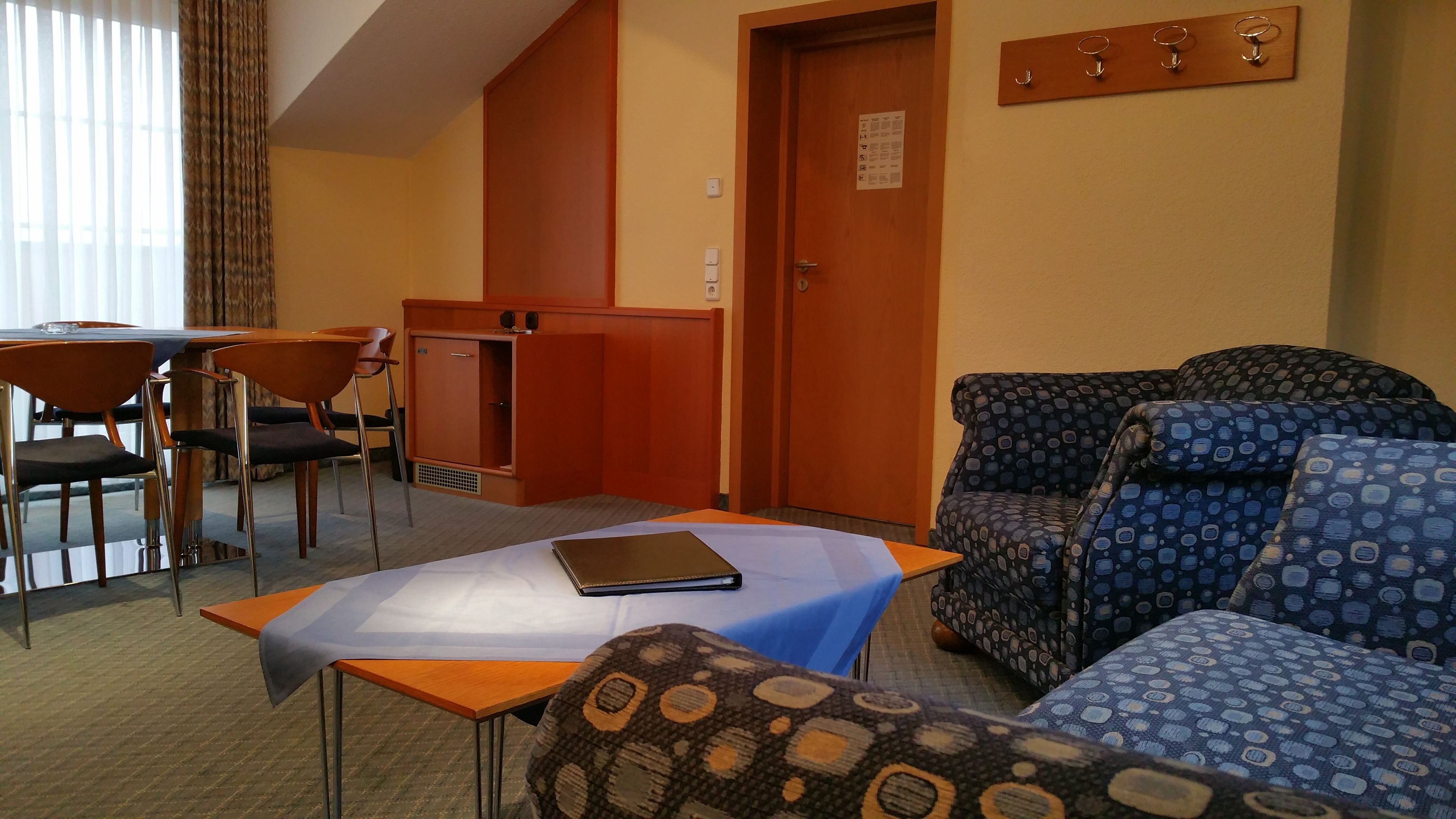 A hotel room with a sofa, a small table with a book, a desk with chairs, and a door with a sign.