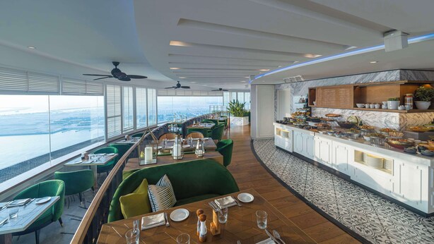 Restaurante moderno com vista alta da cidade, assentos verdes e uma área de buffet.