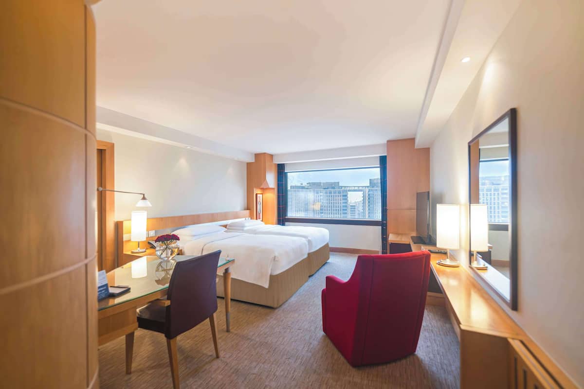 A hotel room with a large bed, a desk with a chair, a red armchair, and a large window offering a city view.