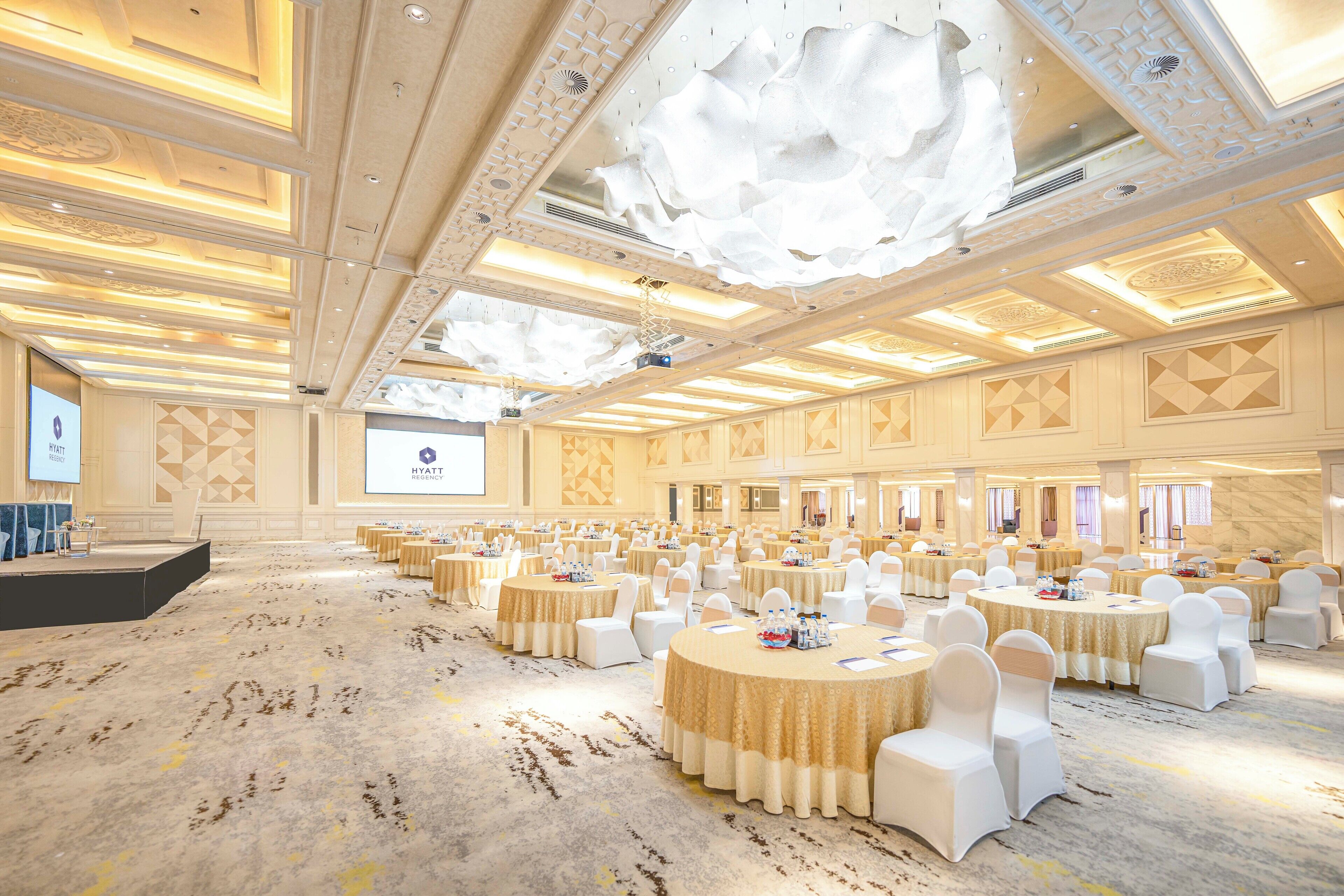Une grande salle de banquet, bien éclairée, avec des tables rondes et des chaises blanches, une scène et un écran de projection.