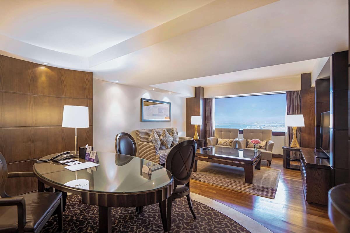 A hotel room with a round glass table, leather chairs, a sofa, and a large window offering a city view.