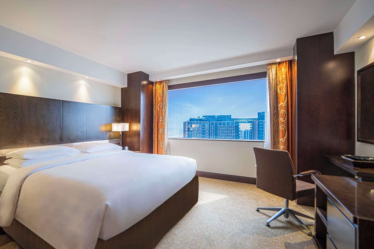 A hotel room with a large bed, a desk, a chair, a TV, and a window with a city view.