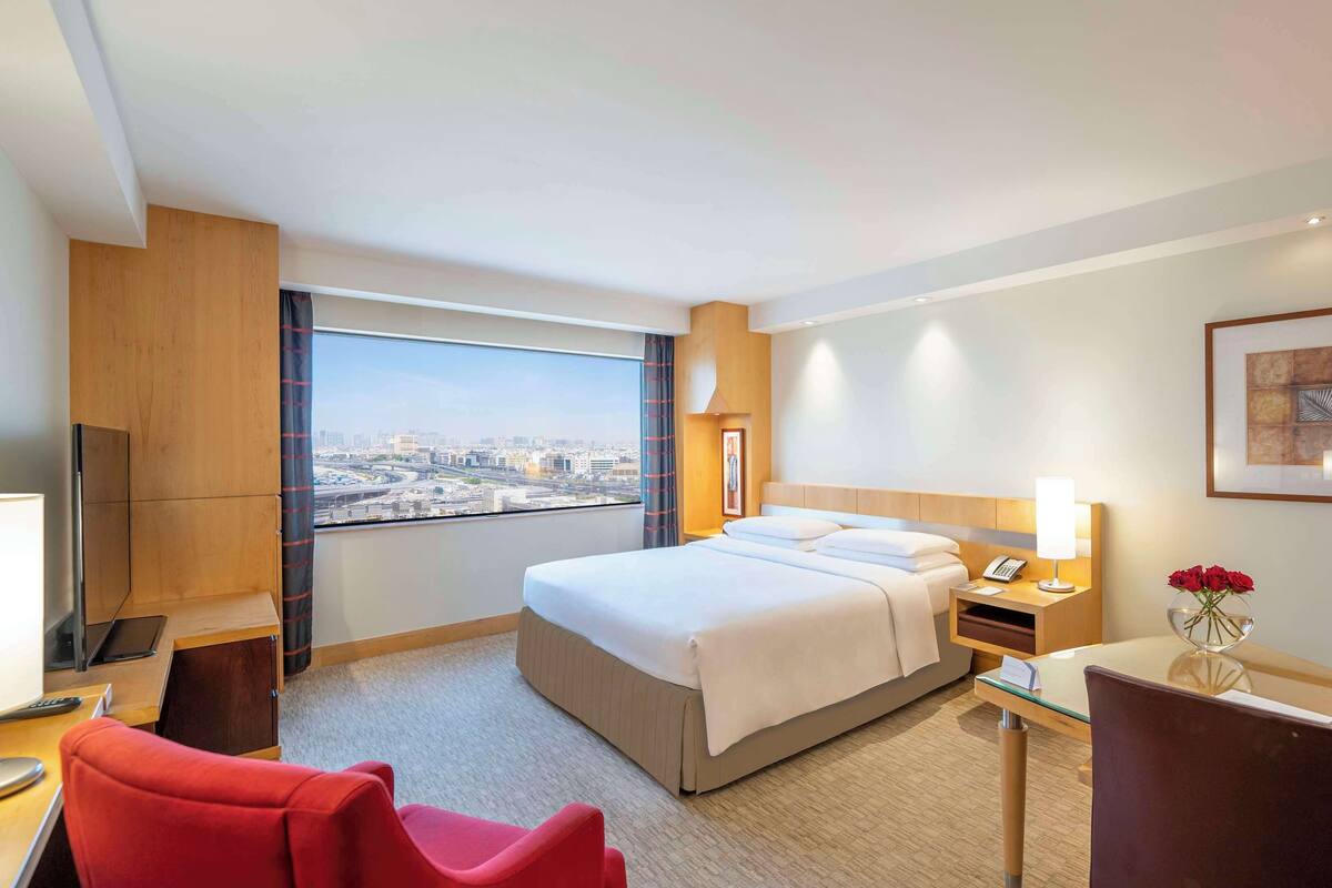 A hotel room with a large bed, a desk with a chair, a TV, a window with a city view, and a framed picture on the wall.