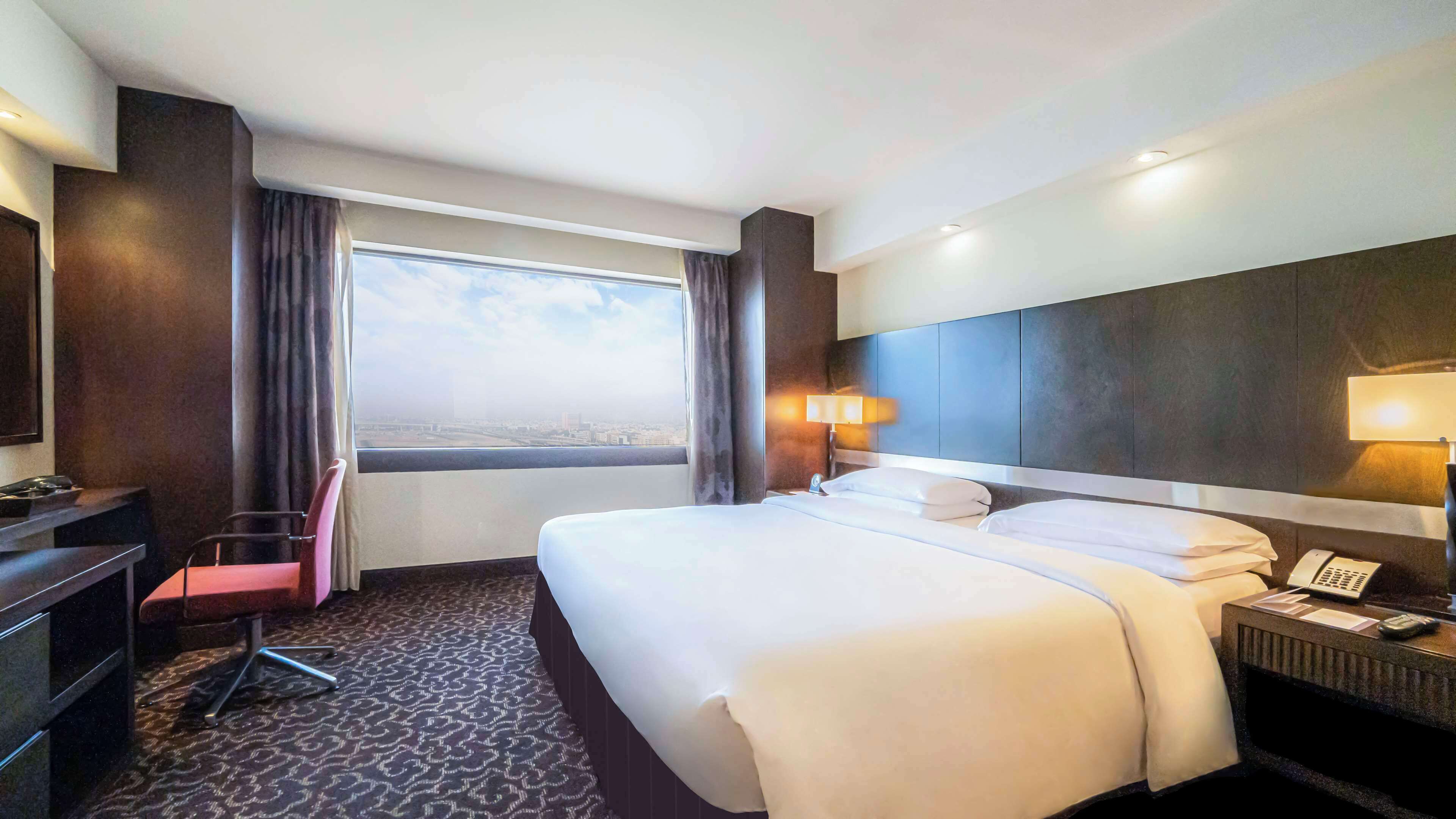Habitación de hotel con una cama grande, un escritorio con silla, una mesita de noche con lámpara y vista a la ciudad desde la ventana.