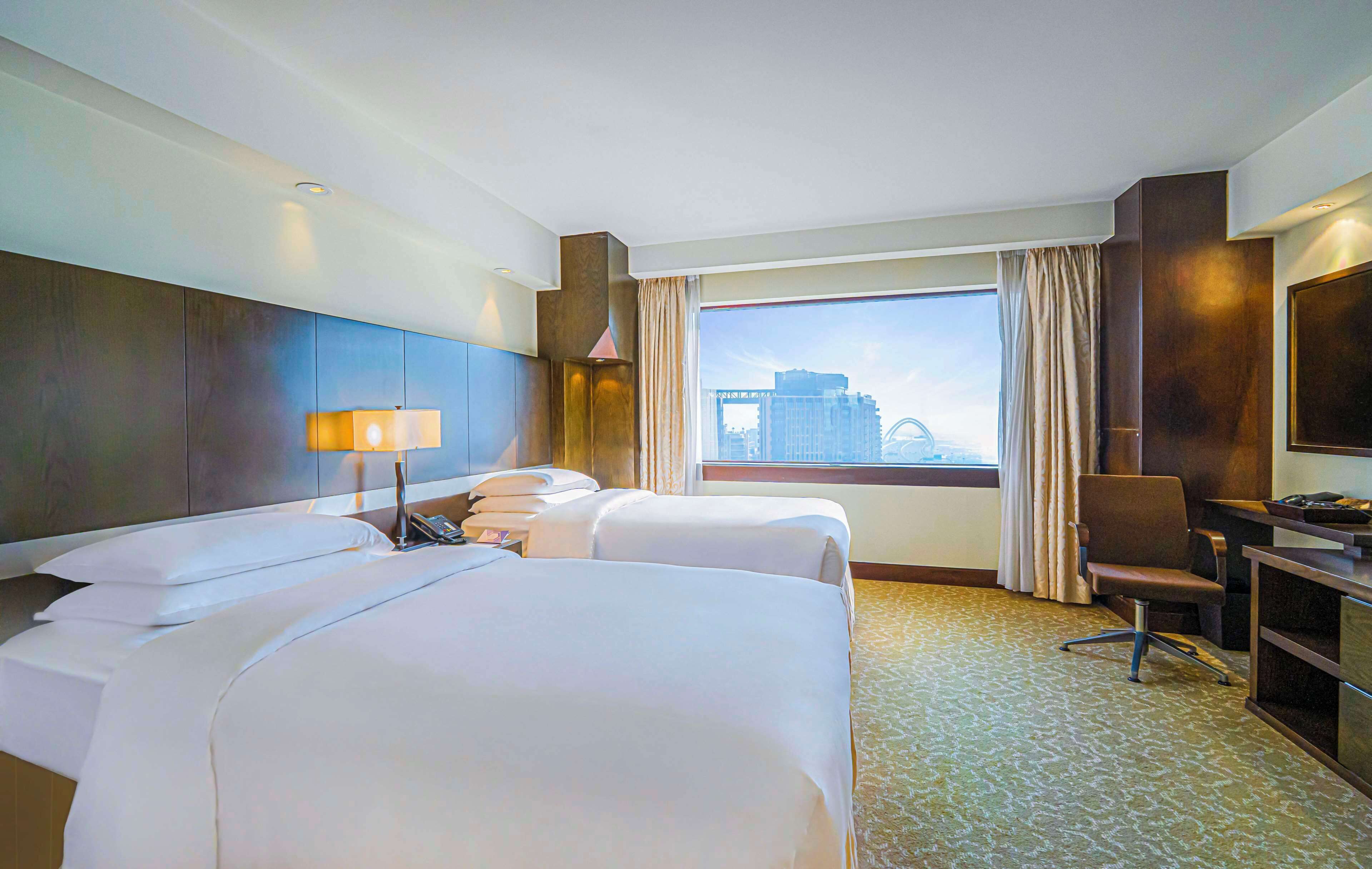 Habitación de hotel con dos camas, un escritorio, una silla, una lámpara y un ventanal con vistas a la ciudad.