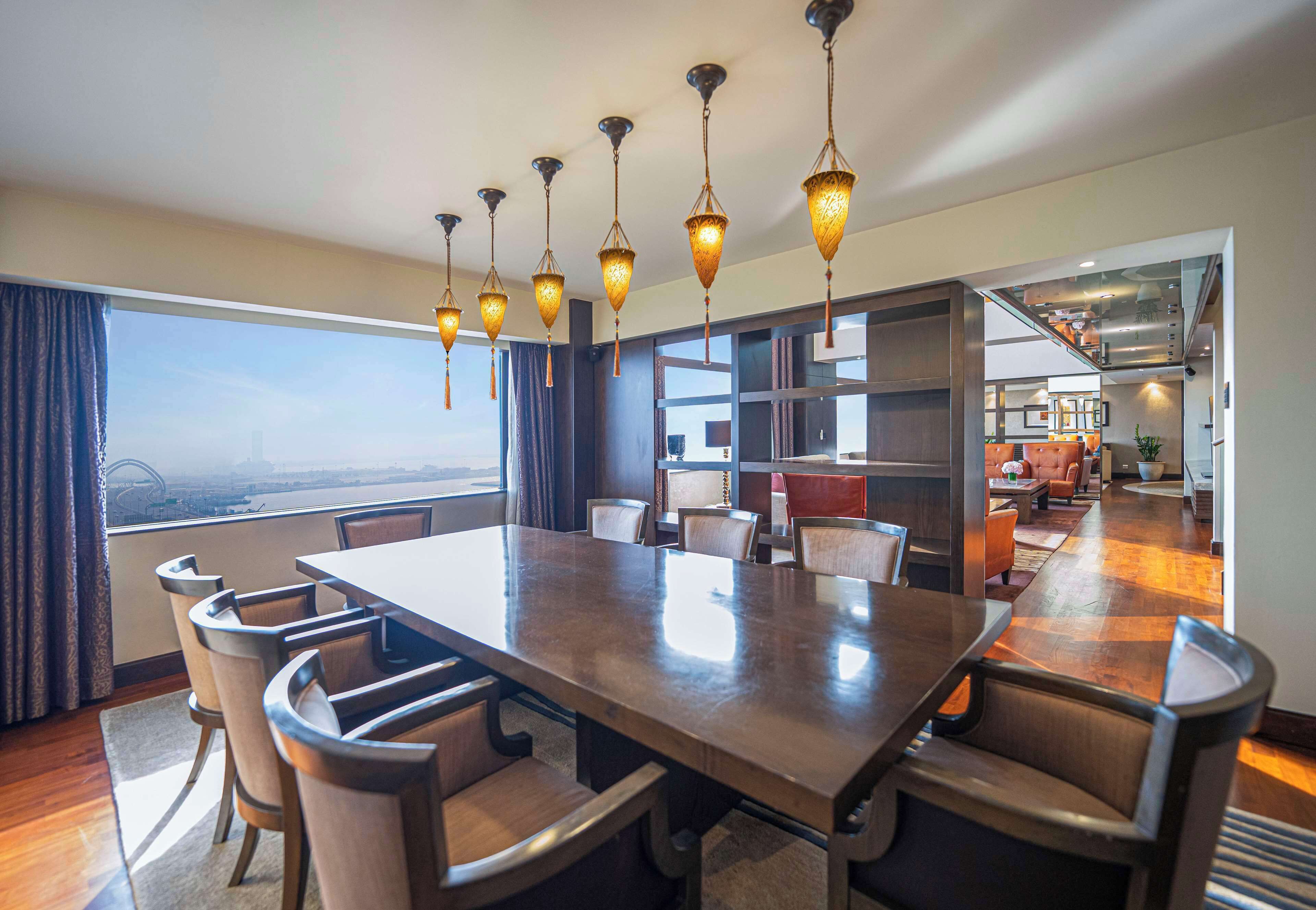 A spacious dining area with a large table and chairs, a view of the city, and a modern kitchen in the background.
