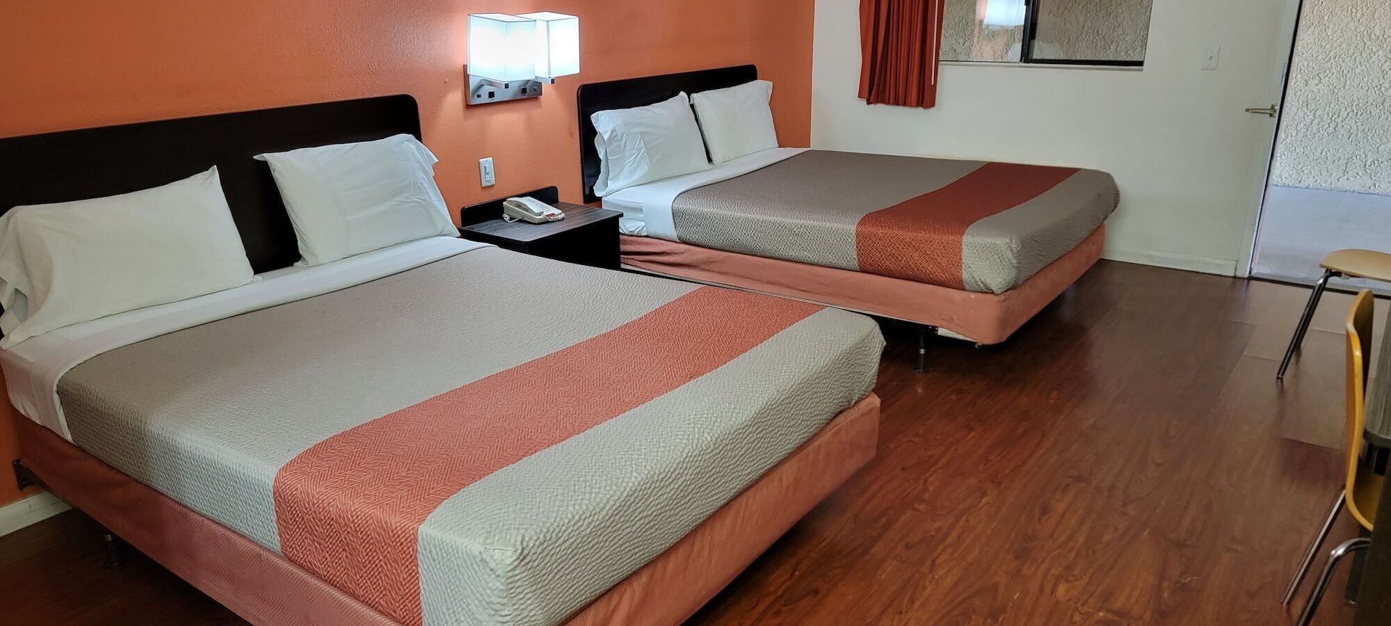A hotel room with two beds, wooden flooring, a nightstand with a phone, and a wall-mounted light.