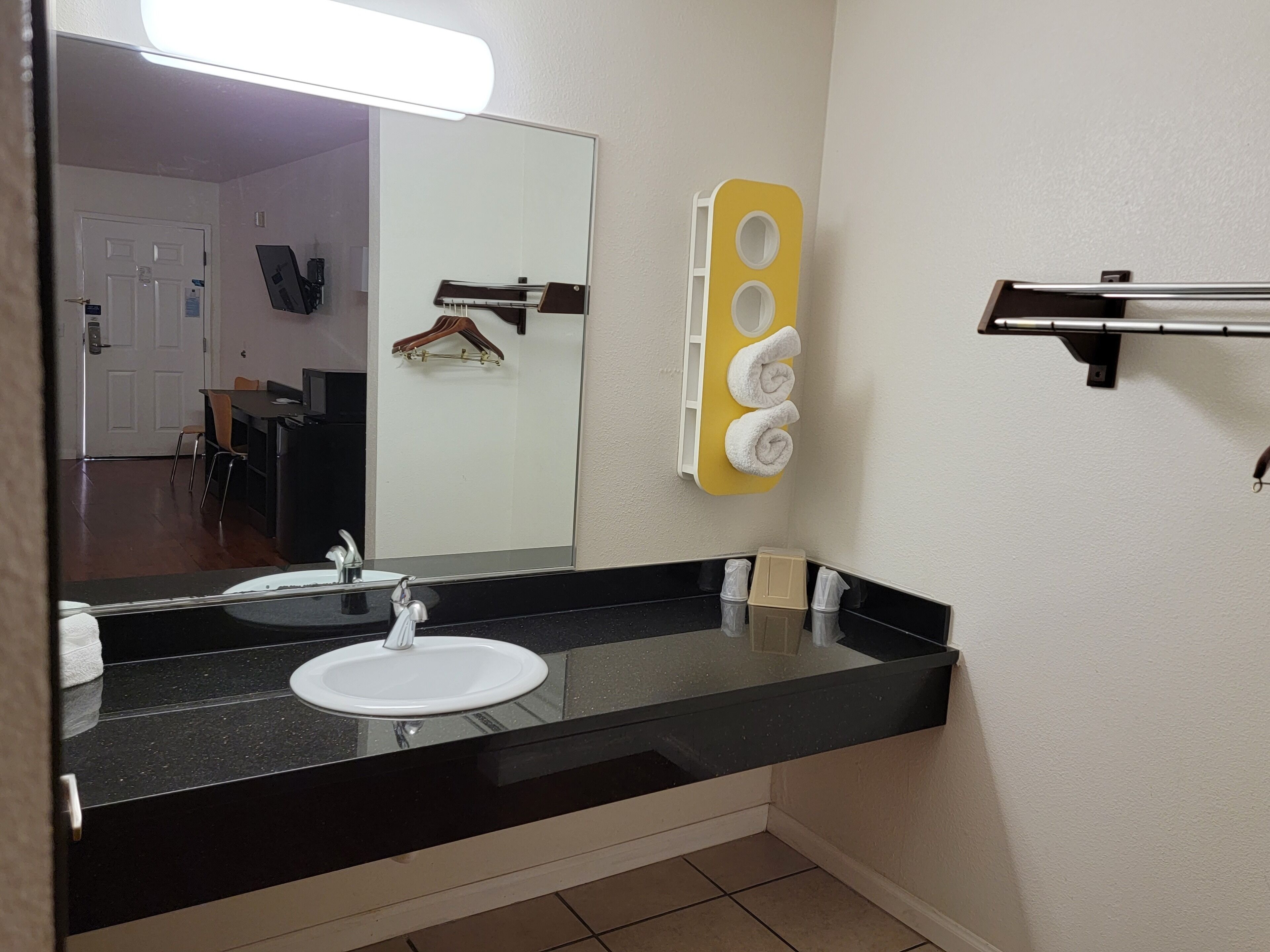 A bathroom with a black countertop, white sink, and a mirror. There is a yellow towel rack with towels and a hanging garment.