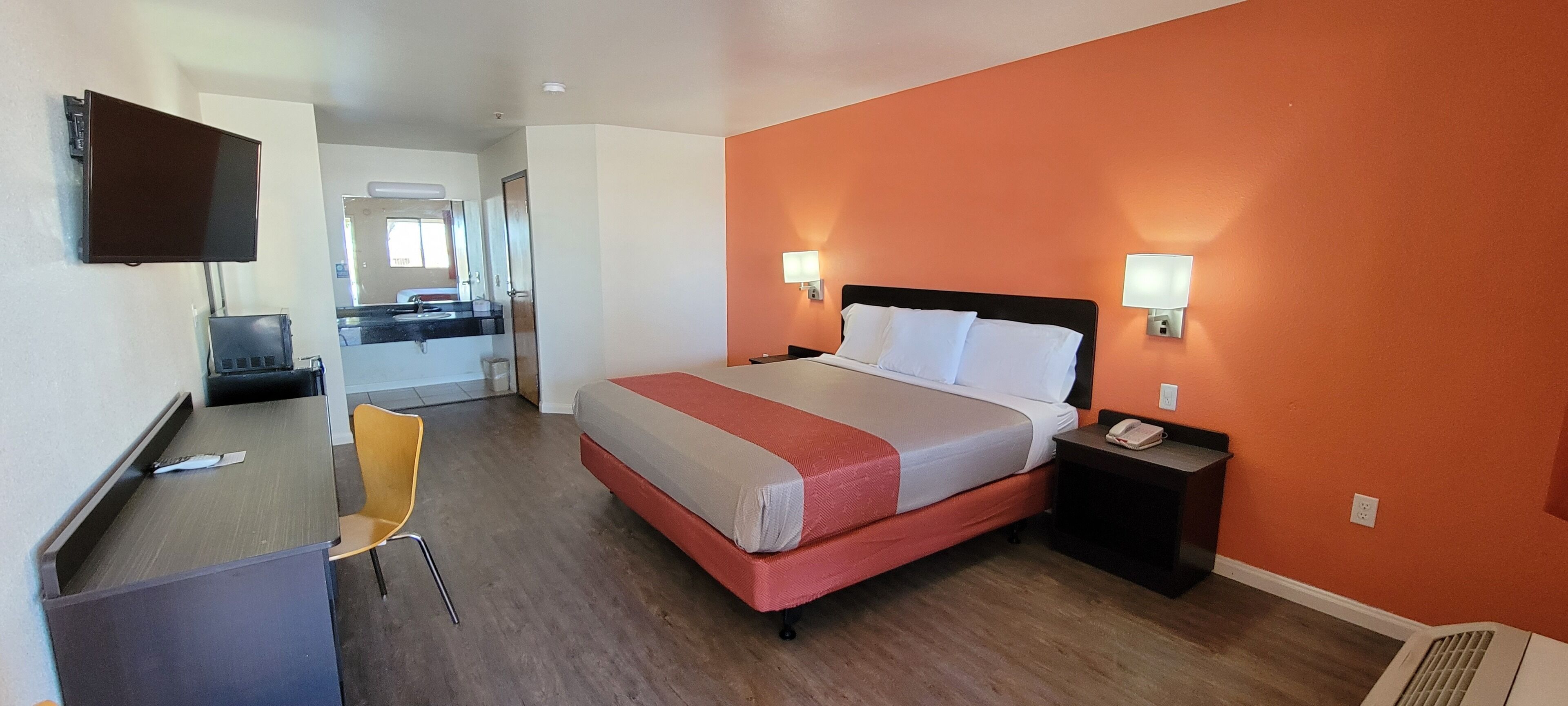Une chambre d’hôtel avec un lit, une table de chevet, une chaise, un bureau, une télévision, et une salle de bain visible au fond.