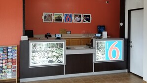 A hotel reception desk with a large illuminated sign displaying the number 6, a check-out counter, a digital screen, and a wall with framed pictures. - Motel 6 Santa Nella, CA - Los Banos (Gustine)