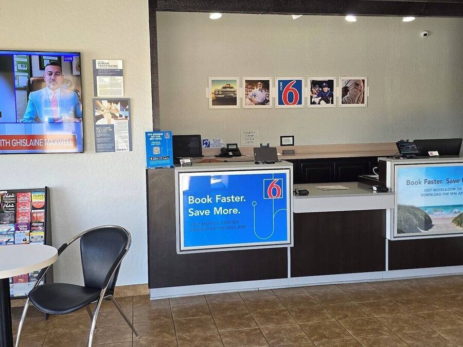 Et hotelreceptionsområde med et fjernsyn der viser et nyhedsprogram, et skrivebord med en computer, en stol, et lille bord og en væg med indrammede billeder samt et skilt, der reklamerer for en mobilapp.