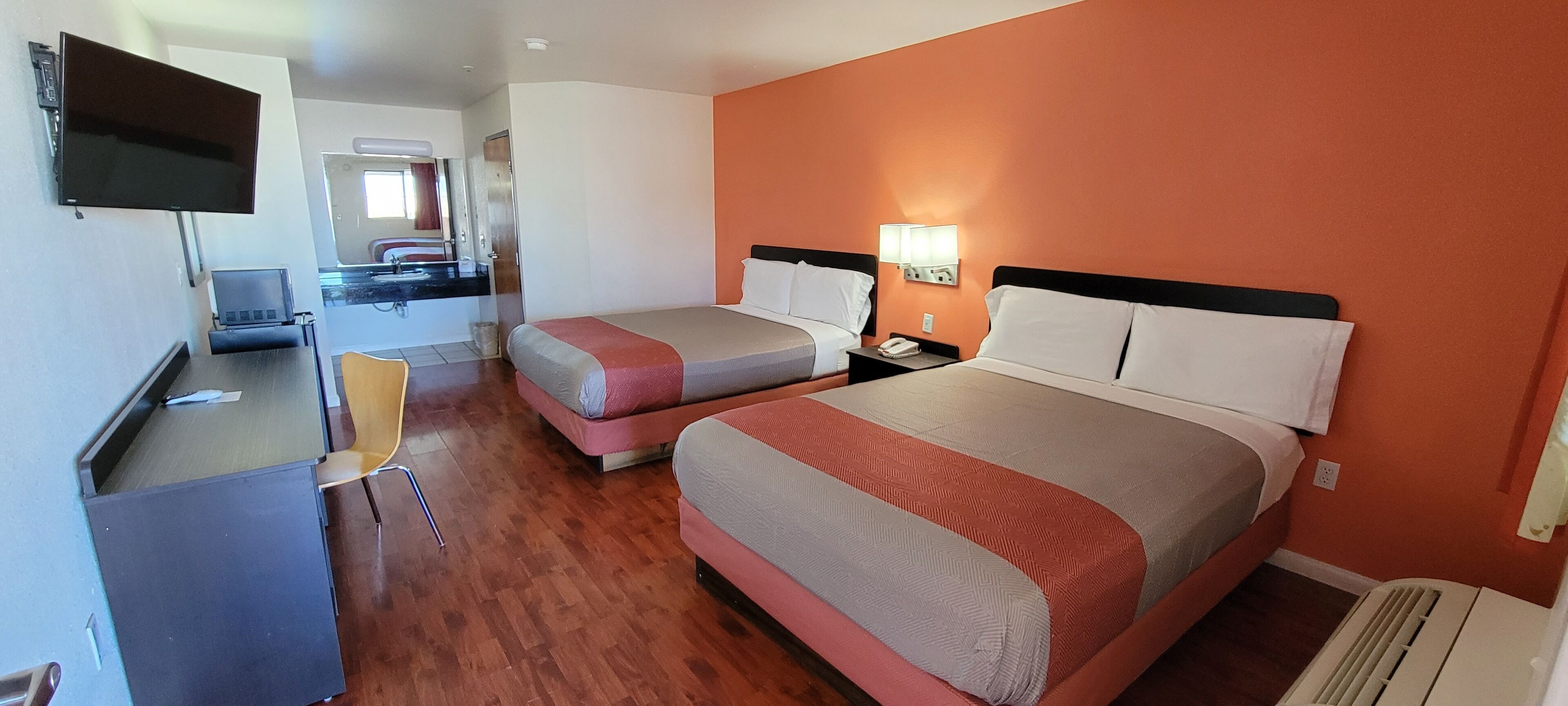 Habitación de hotel con dos camas, un escritorio, un televisor y un baño visible a través de una puerta abierta.
