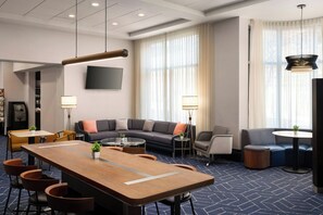 Una habitación de hotel moderna con una gran mesa de madera para comer, un sofá, un televisor y un amplio ventanal con cortinas.