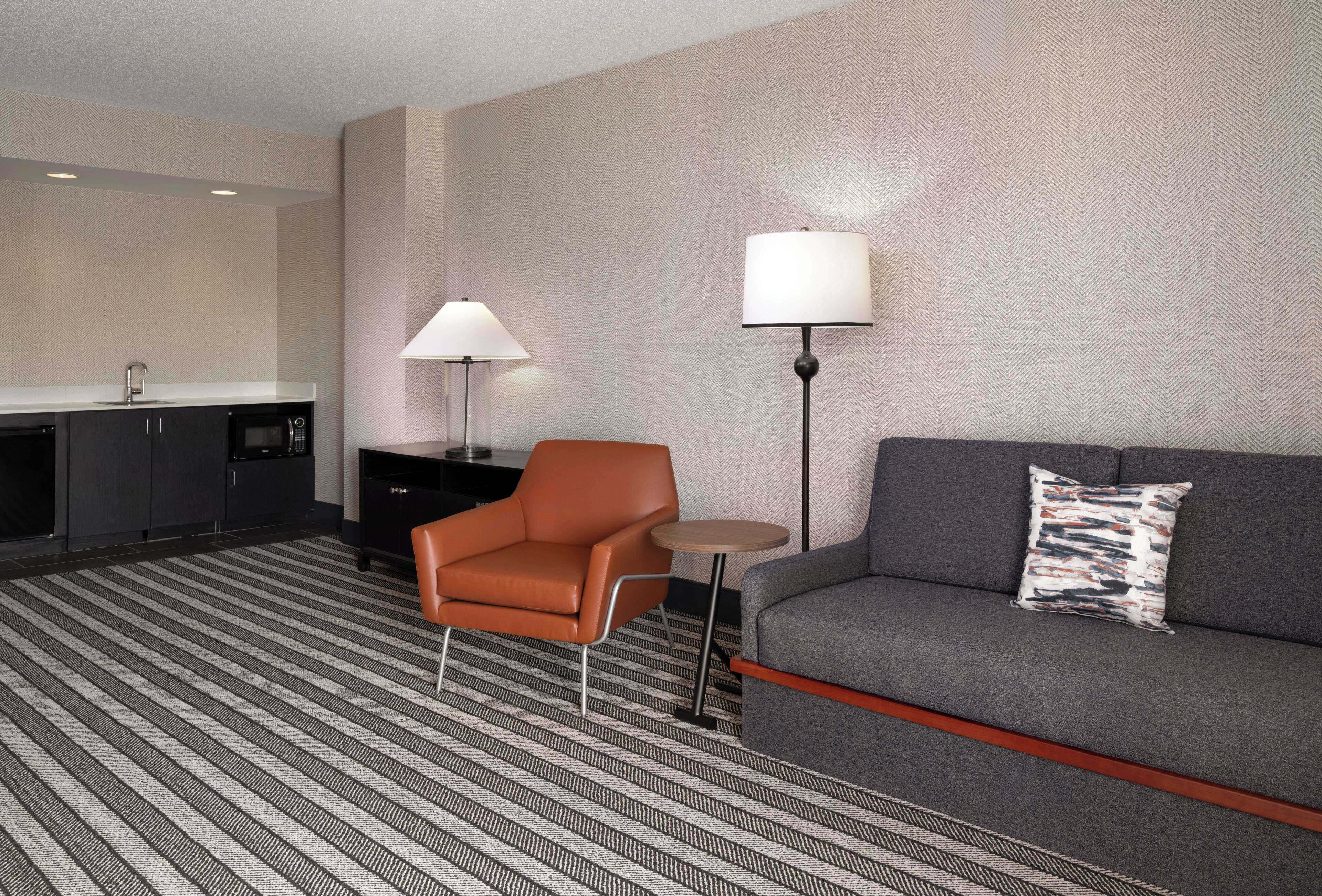 A modern hotel room with a sofa, armchair, small round table, and a lamp.
