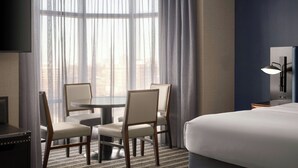A hotel room with a bed, a TV, a dining table with chairs, and a large window with curtains. - Courtyard by Marriott Boston Brookline (Brookline)