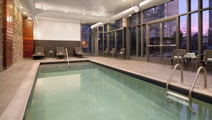 A swimming pool with lounge chairs and a large window with a view of trees. - Courtyard by Marriott Boston Brookline (Brookline)
