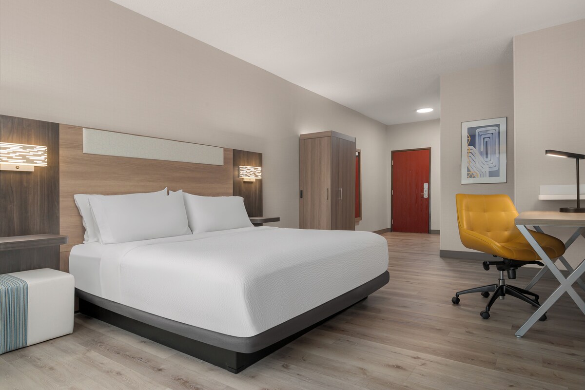 A modern hotel room with a large bed, a desk with a chair, a wardrobe, and a door.