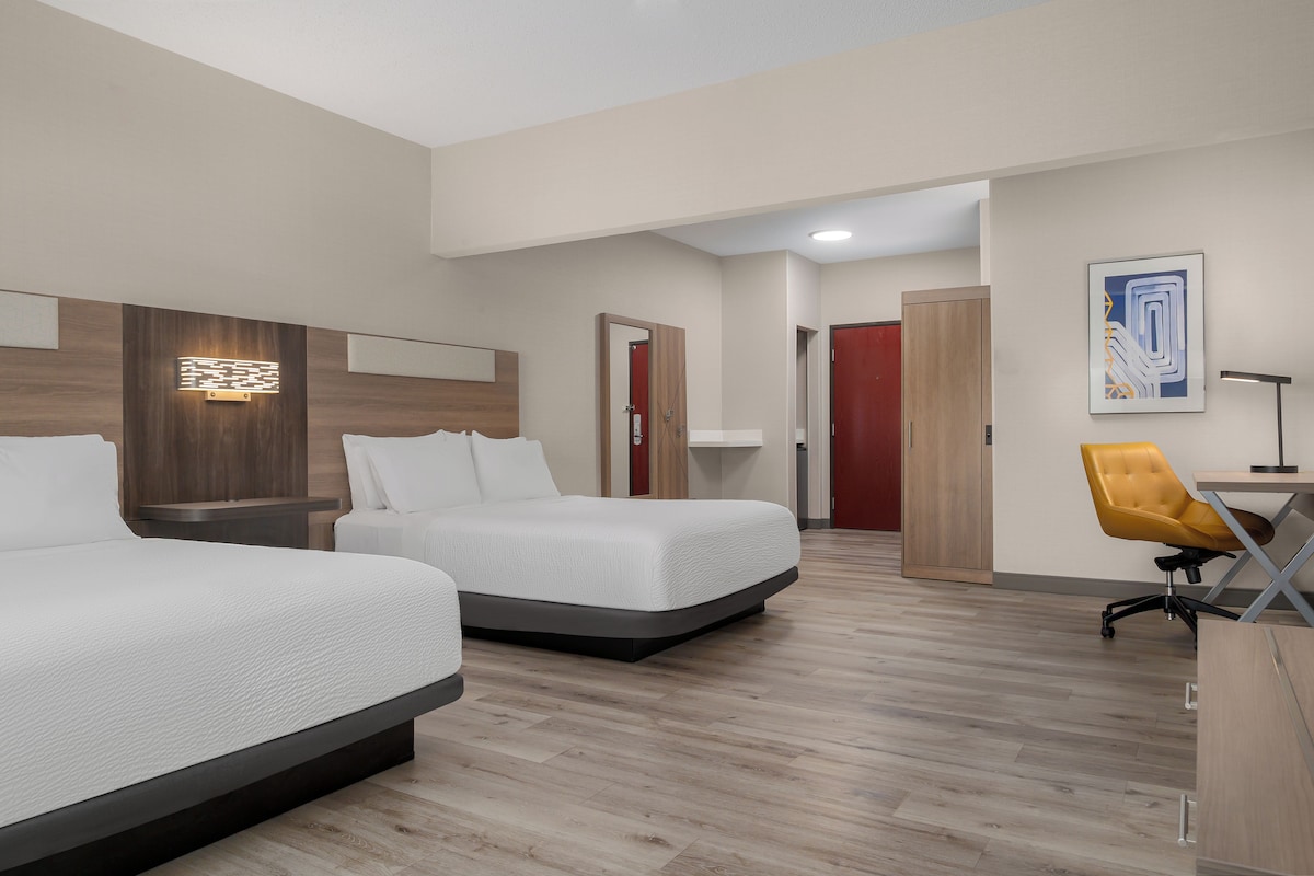 A hotel room with two beds, a desk, a chair, and a door leading to another room.