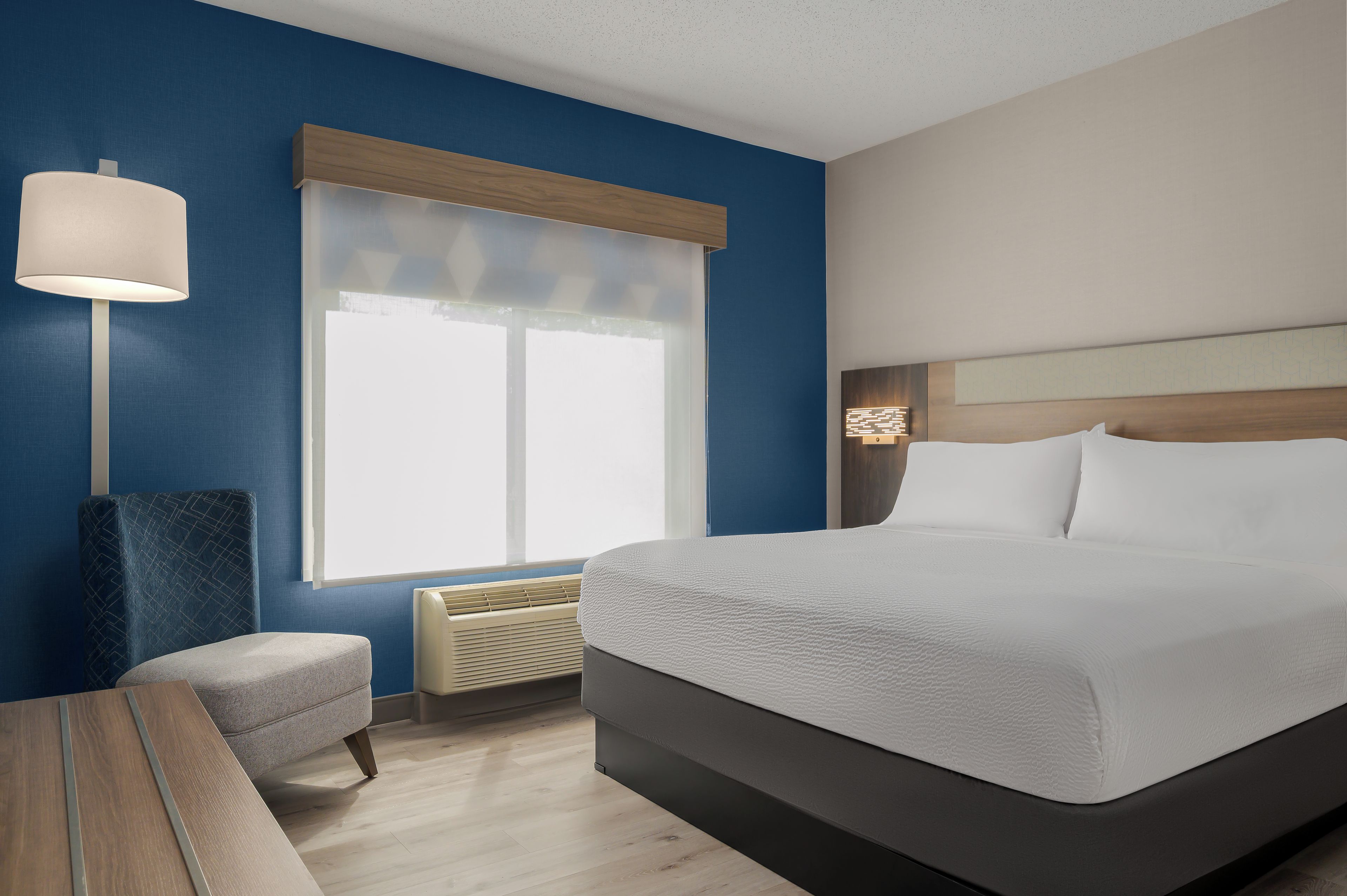 A hotel room with a bed, a bedside table, a lamp, a window with blinds, and a wall-mounted heater.
