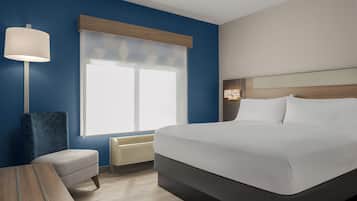A hotel room with a bed, a bedside table, a lamp, a window with blinds, and a wall-mounted heater.