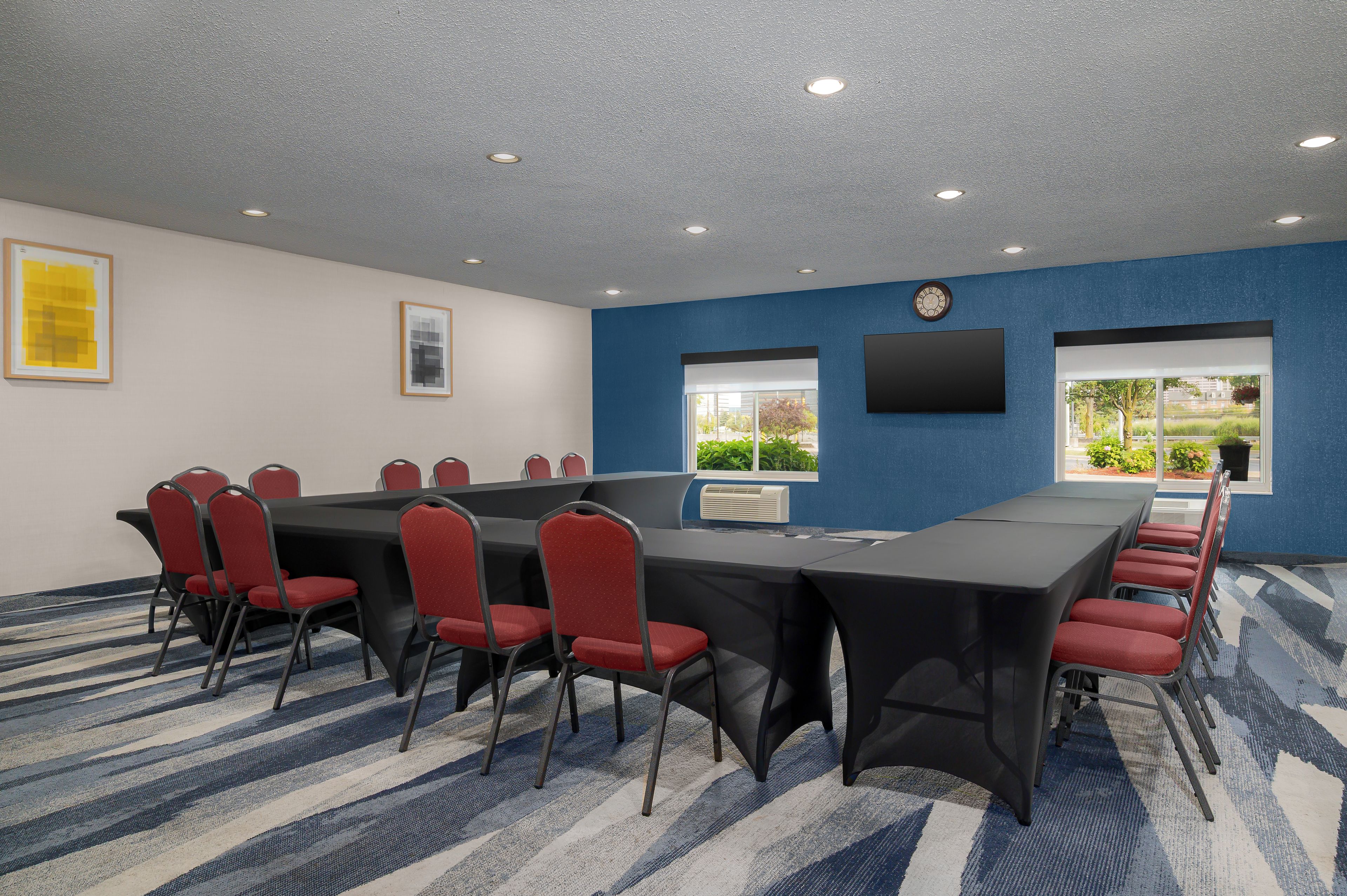 A conference room with a long table, red chairs, a clock, and a window with a view.