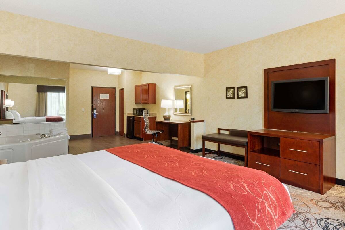A hotel room with a large bed, a desk with a chair, a television, a mirror, and a bathroom.