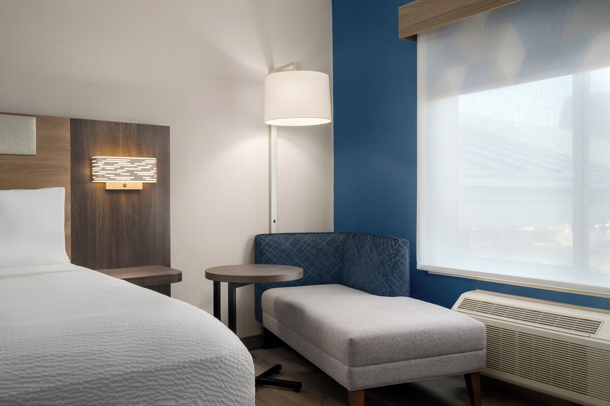 A hotel room with a bed, a small table, a chair, a lamp, and a window.