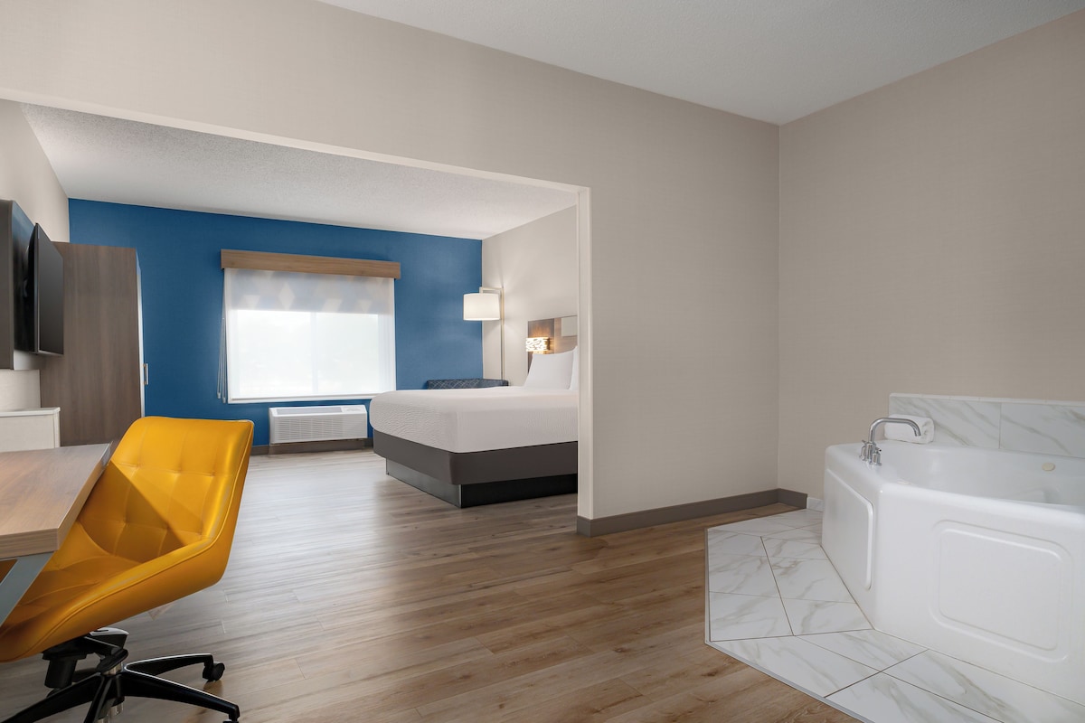 A hotel room with a bed, a desk, a chair, a window, a wall-mounted TV, a lamp, and a bathtub.
