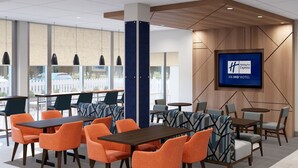 A hotel lobby with a large screen displaying the hotel name, multiple seating areas with tables and chairs, and a view of the outside through large windows. - Holiday Inn Express & Suites Southfield Detroit by IHG (Southfield)