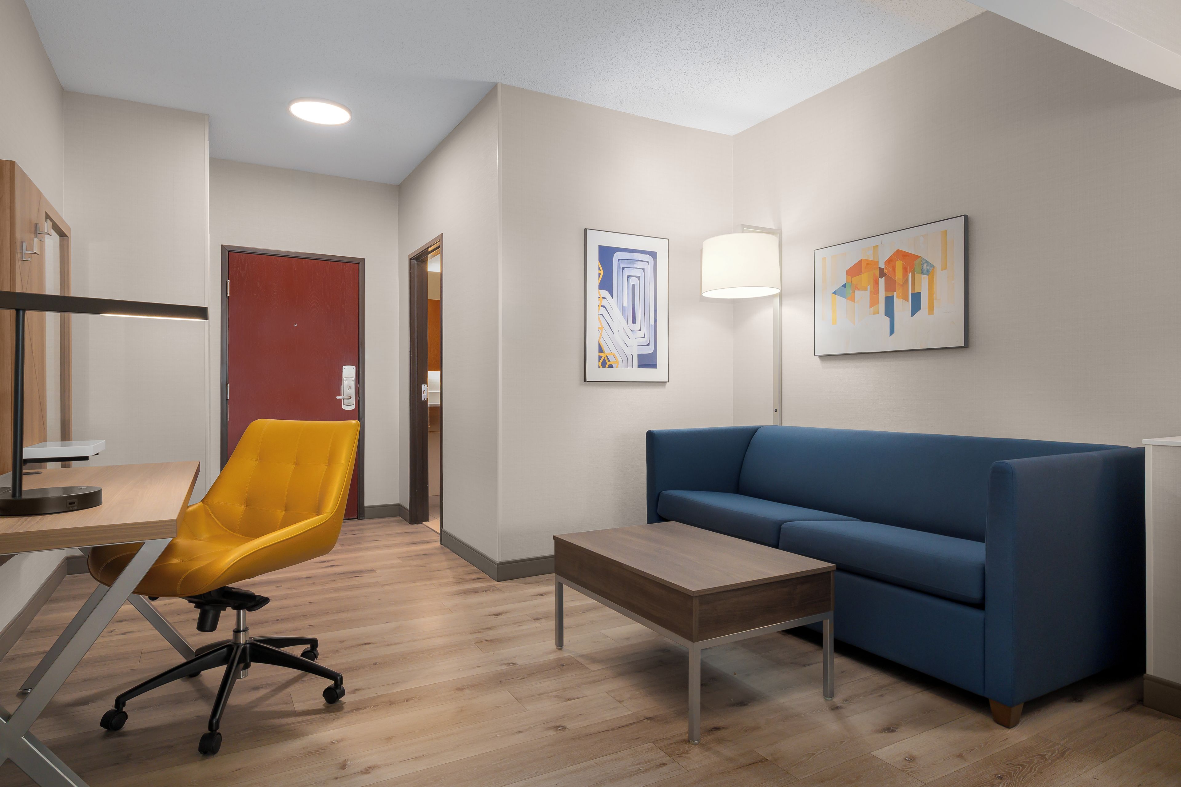 A modern hotel room with a wooden desk, a yellow office chair, a blue sofa, a small table, and two artworks on the wall.