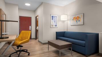 A modern hotel room with a wooden desk, a yellow office chair, a blue sofa, a small table, and two artworks on the wall.